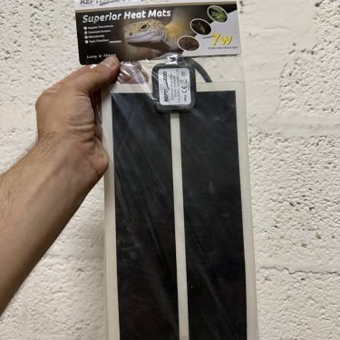 משטח חימום 7W ארוז של REPTI ZOO לזוחלים, מוחזק ביד מול קיר לבנים. אביזר לטרריום, חימום ללטאה.