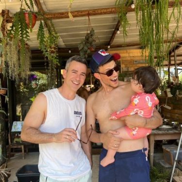זוג גברים שמחים עם תינוקת בחליפת שחייה, מוקפים בצמחייה.