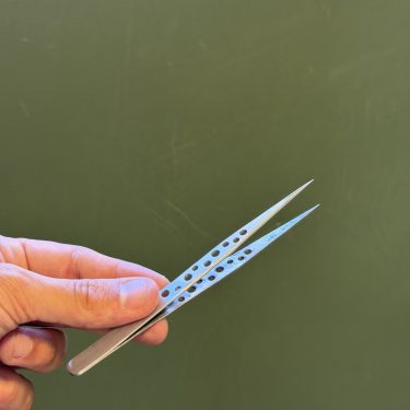 יד אוחזת בפינצטה מחוררת ממתכת על רקע ירוק.